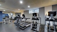 The fitness center at 1303 Delaware Ave in Wilmington, Delaware is a dedicated single-floor exercise space designed to support an active lifestyle. The room features a drop ceiling with recessed lighting that evenly illuminates the space, complemented by a horizontal window along one wall that allows natural light to filter in. The flooring consists of durable rubber gym matting, providing both safety and comfort during workouts. A large wall mirror expands the sense of space and allows users to monitor their form during exercise. The facility is equipped with True-brand cardio machines, including multiple treadmills and an elliptical trainer, reflecting a commitment to quality fitness equipment. Additional strength and conditioning equipment is visible toward the rear of the space. The overall aesthetic is clean and functional, with a neutral color palette of grays and whites contributing to a focused workout environment. The layout is thoughtfully arranged to maximize usable floor space while maintaining comfortable circulation between equipment. This well-maintained fitness center offers residents a convenient and professional-grade exercise facility on-site.