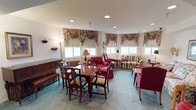 This single-floor communal space is thoughtfully designed to foster comfort and social interaction, blending classic decor with a warm, residential ambiance. The main lounge area is generously proportioned, featuring large windows dressed with elegant floral swag valances that allow an abundance of natural light to fill the room. The curved bay of windows creates an inviting seating nook furnished with plaid and floral-upholstered sofas and armchairs, complemented by side tables and floor lamps that add a soft, homey glow. A traditional wooden dining table surrounded by Chippendale-style chairs serves as a central gathering point, ideal for games, activities, or shared meals. An upright piano anchors one corner, adding a touch of classic charm and providing a focal point for musical engagement. Framed artwork adorns the walls, contributing to the refined, traditional aesthetic throughout. An adjacent activity or reading room offers a more relaxed setting, with a daybed, bookshelf stocked with reading materials, and cozy seating. A large-screen television housed in a dark wood entertainment unit provides additional recreational options. The teal-patterned carpeting runs throughout, tying the spaces together with a cohesive, classic feel. French glass doors connect the areas, enhancing the open and airy character of the overall space.
