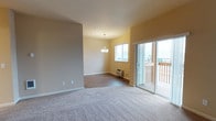 This single-floor space features an open-concept layout that combines a living area and dining space in a seamless, functional design. The living area is finished with plush, neutral-toned carpeting, while the adjoining dining area transitions to wood-look hard flooring, creating a subtle distinction between the two zones. Warm, light-toned walls in soft beige and yellow hues contribute to an inviting atmosphere throughout.

The dining area is highlighted by a modest chandelier that serves as a central decorative element, complemented by a standard window that allows natural light to filter into the space. A wall-mounted heating unit is positioned beneath the window, providing a practical climate-control solution. 

A sliding glass door connects the interior to an outdoor deck, enhancing the sense of space and bringing in additional natural light. The deck features a wooden railing and offers views of the surrounding neighborhood. White baseboards run throughout, providing a clean finish against the warm wall tones. Overall, the space presents a straightforward, comfortable layout suited for everyday living.