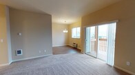 This single-floor space features an open-concept layout that combines a living area and dining space in a seamless, functional design. The living area is finished with plush, neutral-toned carpeting, while the adjoining dining area transitions to wood-look hard flooring, creating a subtle distinction between the two zones. Warm, light-toned walls in soft beige and yellow hues contribute to an inviting atmosphere throughout.

The dining area is highlighted by a modest chandelier that serves as a central decorative element, complemented by a standard window that allows natural light to filter into the space. A wall-mounted heating unit is positioned beneath the window, providing a practical climate-control solution. 

A sliding glass door connects the interior to an outdoor deck, enhancing the sense of space and bringing in additional natural light. The deck features a wooden railing and offers views of the surrounding neighborhood. White baseboards run throughout, providing a clean finish against the warm wall tones. Overall, the space presents a straightforward, comfortable layout suited for everyday living.