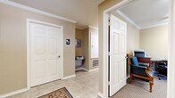 This single-floor community space offers a thoughtfully organized layout designed to serve multiple functions comfortably. The combined dining and living area forms the heart of the space, featuring warm beige walls, crown molding, and plush carpeted flooring throughout. The living area is furnished with a generous curved sectional sofa, ceiling fans, and fluorescent lighting, while large windows dressed with decorative valances and horizontal blinds allow natural light to fill the room and provide views of the surrounding outdoor greenery. The adjacent dining area accommodates multiple round and rectangular tables with upholstered chairs, making it well-suited for group gatherings. Colorful wall décor adds a lively touch to the room's welcoming atmosphere. The business center office is a dedicated, functional workspace equipped with computer workstations, a printer, and ample desk space. A posted rules board and window with draped curtains complete the professional yet approachable setting. The hallway connects the key areas of the space and includes an accessible restroom, a water fountain, and a decorative area rug over tile flooring, with crisp white six-panel doors and crown molding maintaining a cohesive, polished aesthetic throughout.