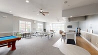 Located at 6336 Clock Tower Dr in Walbridge, Ohio, this generously sized single-floor game room serves as a vibrant community gathering space. The room is thoughtfully designed with a neutral gray and white color palette, wainscoting along the lower walls, and a combination of plush carpeting and tile flooring throughout. Recessed lighting and dual ceiling fans ensure the space is well-lit and comfortable year-round. Large multi-pane windows along multiple walls flood the interior with abundant natural light, while glass doors provide direct access to an outdoor pool area, creating a seamless indoor-outdoor connection. The game room is well-equipped for entertainment, featuring a ping-pong table, a foosball table, and a billiards table. A dedicated lounge area offers comfortable leather seating and a wall-mounted television, ideal for relaxing between activities. A bar-style counter with barstool seating adds a social hub to the space, complemented by a small work or dining table nearby. The open layout allows for easy movement between activity zones, making this a versatile and welcoming venue for residents to gather, play, and unwind.