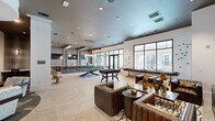 Located at 1515 Rollick Rd in Cary, NC, this single-floor community amenity space offers a generously sized, thoughtfully designed environment ideal for socializing and relaxation. The open-concept main living area features large floor-to-ceiling windows that flood the space with natural light and offer views of the surrounding community and pool area. Polished stone tile flooring, recessed lighting, and pendant fixtures contribute to the modern, upscale aesthetic throughout. A dedicated game room includes a pool table and foosball table, set beneath dramatic pendant lighting, making it a standout entertainment hub. Rich leather seating arrangements, layered rugs, and curated artwork create a sophisticated lounge atmosphere in the adjoining living area. A separate lounge zone features a flat-screen TV mounted on a textured accent wall, complemented by plush seating and bold patterned rugs. The covered outdoor terrace is equipped with ceiling fans, a linear gas fireplace set into a stacked stone wall, a wall-mounted TV, and outdoor lounge furniture on turf accents, offering a comfortable year-round retreat with views of the resort-style pool. Inside, intimate wood-paneled booth seating and a boldly patterned carpet define a lounge and co-working nook, adding a warm contrast to the brighter open areas. The overall ambiance is contemporary, inviting, and versatile.