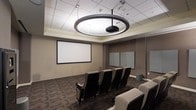 This single-floor presentation and screening room is thoughtfully designed to facilitate focused viewing and collaborative sessions. The space is anchored by a large wall-mounted projection screen at the front, paired with a ceiling-mounted projector suspended from a striking circular ring light fixture that serves as both a functional and architectural centerpiece. The recessed ceiling detail surrounding this fixture adds a layered, polished look to the room's overhead design.

Rows of theater-style seats with padded upholstery and armrests are arranged to face the screen, providing a comfortable and organized viewing experience for attendees. The neutral, taupe-toned walls complement the dark patterned carpeting, creating a professional and distraction-free ambiance. Along one side wall, a set of whiteboards offers additional space for note-taking or brainstorming during presentations. The drop ceiling with recessed panel tiles contributes to the room's acoustic and visual cohesion. Overall, this space is well-suited for corporate presentations, media screenings, or training sessions.