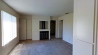 This single-floor residence offers a straightforward and functional layout designed for comfortable everyday living. The main living area is finished with wall-to-wall carpet and neutral-toned walls, creating a clean and versatile backdrop suited to a variety of interior styles. A standout feature of the space is the built-in fireplace, framed by a white surround and set into a central wall, serving as a natural focal point for the room. Adjacent to the fireplace, built-in shelving provides convenient storage and display options. A ceiling fan is installed overhead, contributing to year-round climate comfort. The living area benefits from natural light streaming through a window fitted with vertical blinds, adding brightness to the space. HVAC floor vents are present throughout, ensuring efficient heating and cooling. A series of closet doors along one wall offer generous storage capacity. The layout flows between areas with open doorways connecting the main living space to additional rooms beyond. The overall ambiance is calm and neutral, presenting a well-maintained, move-in-ready environment with solid functional bones and ample opportunity for personalization.