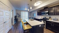 This single-floor residence is designed with a modern, cohesive aesthetic highlighted by dark espresso cabinetry, white countertops, and wood-style flooring that runs throughout the main living areas. The kitchen is a standout feature, offering a large island with a built-in double sink, pendant lighting, and a full suite of stainless steel appliances including a gas range, microwave, and refrigerator. Ample cabinetry provides generous storage, while the island doubles as a casual dining space with seating for multiple guests. The kitchen flows naturally into the living area, which benefits from a sliding glass door fitted with vertical blinds, allowing natural light to filter in. The bedroom is carpeted for comfort, features a sizable window with blinds, and connects conveniently to the rest of the unit. The bathroom is clean and functional, equipped with dark vanity cabinetry, a white countertop, a tub-shower combination, and wood-style flooring that ties into the overall design. Throughout, the neutral gray walls and consistent finishes create a unified, contemporary ambiance.