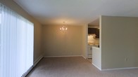 This single-floor residence offers a thoughtfully arranged layout with a seamless connection between the main living and dining area and the adjacent kitchen. The dining space is anchored by an elegant candle-style chandelier that adds a warm, classic touch to the room. Neutral beige walls and plush wall-to-wall carpeting create a cozy and inviting atmosphere throughout. A baseboard heating unit runs along the lower wall, ensuring comfort year-round, while a large window allows natural light to fill the space. The kitchen is partially open to the dining area via a pass-through counter, making it ideal for entertaining and everyday living. It features dark espresso-toned cabinetry, a white range with overhead range hood, and light-colored countertops that provide a clean contrast to the darker upper cabinets. The overall design is understated and functional, with white trim accents framing the walls and doorways to give the space a tidy, finished look. The open sightlines between rooms contribute to an airy, connected feel across the entire floor plan.