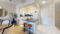 This single-floor residence offers a thoughtfully designed open-concept layout that seamlessly connects the kitchen, dining, and living areas. The kitchen is outfitted with white cabinetry, white appliances, and a peninsula island topped with a dark countertop, accommodating bar stools for casual dining. Tile flooring transitions into carpet as the space opens into the combined dining and living area.

The living room is a welcoming, light-filled space anchored by a bold teal accent wall and a ceiling fan overhead. A sliding glass door leads to an outdoor balcony, filling the room with natural light. Built-in shelving adds functional display space, and the layout comfortably accommodates both a seating area and a dining table with chairs.

The bedroom features plush carpeting, a natural light source through a draped window, and recessed lighting. Ample space allows for a full furniture arrangement including a dresser, seating, and bedside tables.

The bathroom is finished with white tile flooring, an extended vanity with white cabinetry, and a tub-shower combination. An adjacent walk-in closet provides generous storage. Throughout the unit, a neutral color palette creates a clean, cohesive ambiance.