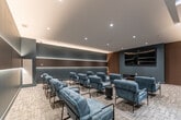 Located at 375 South St in London, Ontario, this private screening room is a thoughtfully designed amenity space that delivers a refined cinematic experience. The room is anchored by a large wall-mounted display accompanied by a low-profile media console, set against a striking accent wall finished in warm wood slat paneling and cool slate-blue tones. Rows of plush, blue-grey upholstered lounge chairs with sleek black metal frames are arranged in a theatre-style layout, providing generous seating for a group. Each seating area is complemented by compact side tables, enhancing comfort and functionality. The back and side walls feature a distinctive horizontal band of backlit dark wood paneling, casting a warm ambient glow that adds depth and drama to the space. Recessed ceiling lighting provides clean, even illumination throughout, while a coffered ceiling detail lends an additional layer of architectural refinement. The floor is finished in a neutral, textured carpet tile that ties the contemporary palette together. A built-in shelving and cabinetry unit on one wall provides additional storage and display space. The overall ambiance is polished and intimate, combining modern design sensibilities with a welcoming atmosphere ideal for private screenings or group gatherings.