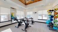 Located at 1003 148th St W in Rosemount, MN, this dedicated exercise studio offers a well-appointed space designed for group fitness and cycling workouts. The room is defined by its striking vaulted ceiling, clad in warm wood paneling and fitted with a large ceiling fan, which adds both visual character and ventilation. Recessed lighting throughout ensures the space is evenly and brightly lit at all times.

Large windows with roller shades line the walls, inviting generous natural light and providing views of the surrounding neighborhood. The neutral, textured carpet flooring offers a comfortable surface underfoot, while clean white walls contribute to an open, energizing atmosphere.

The exercise area is equipped with multiple stationary spin bikes arranged to face the windows and a wall-mounted flat-screen TV, supporting both instructor-led and self-guided workouts. A full-length mirror spans one wall, enhancing the sense of space and allowing for form monitoring. A well-stocked equipment rack holds an assortment of medicine balls, resistance bands, yoga mats, and kettlebells, making the studio versatile for a variety of fitness routines.

Entry is provided through elegant double glass-paned doors, and an interactive fitness mirror station is positioned near the entrance. This thoughtfully designed studio delivers a focused, energizing environment suited for cycling, yoga, and group exercise classes.