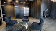 This single-floor space presents a striking modern lobby with a carefully curated design aesthetic that blends industrial and contemporary elements. The interior is defined by dramatic dark wall paneling with subtle horizontal striping, lending depth and a sense of refined sophistication to the space. Light hardwood flooring provides a warm contrast against the predominantly dark palette, grounding the space and enhancing its visual balance.

A stainless steel elevator serves as a functional focal point along the main wall, seamlessly integrated into the design with a clean, architectural surround. Built-in shelving units flank the elevator, adding both structure and utility to the space.

The seating arrangement is thoughtfully composed, featuring a collection of plush, dark velvet accent chairs positioned around a sculptural glass-top coffee table with a distinctive metallic base. The arrangement encourages a comfortable yet stylish gathering area, suitable for welcoming visitors or facilitating informal interactions.

The overall ambiance is polished and professional, with a cohesive dark color scheme punctuated by metallic accents throughout. The space conveys a strong sense of design intentionality, making it well-suited for a high-end commercial, hospitality, or mixed-use environment.
