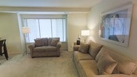 This single-floor space features a well-appointed living area designed with comfort and functionality in mind. The lounge is finished with plush, neutral-toned carpeting that adds warmth underfoot and contributes to the overall soft, inviting atmosphere of the room. Two generously sized sofas in a taupe/mocha upholstery are arranged to encourage conversation and relaxation, each accented with coordinating throw pillows.

A wide window dressed with horizontal blinds allows natural light to filter into the space, brightening the neutral beige walls and creating an airy feel. A side table positioned between the seating area holds a classic table lamp that provides warm ambient lighting, complemented by a decorative ceramic vase. A framed piece of abstract artwork adorns the wall, adding a subtle splash of color to the otherwise understated palette.

The ceiling features a recessed tray detail, lending a modest architectural element to the room. Overall, the space presents a clean, cohesive aesthetic with a calm and comfortable ambiance suited for everyday living.