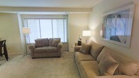 This single-floor space features a well-appointed living area designed with comfort and functionality in mind. The lounge is finished with plush, neutral-toned carpeting that adds warmth underfoot and contributes to the overall soft, inviting atmosphere of the room. Two generously sized sofas in a taupe/mocha upholstery are arranged to encourage conversation and relaxation, each accented with coordinating throw pillows.

A wide window dressed with horizontal blinds allows natural light to filter into the space, brightening the neutral beige walls and creating an airy feel. A side table positioned between the seating area holds a classic table lamp that provides warm ambient lighting, complemented by a decorative ceramic vase. A framed piece of abstract artwork adorns the wall, adding a subtle splash of color to the otherwise understated palette.

The ceiling features a recessed tray detail, lending a modest architectural element to the room. Overall, the space presents a clean, cohesive aesthetic with a calm and comfortable ambiance suited for everyday living.