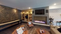 This thoughtfully designed single-floor space blends modern aesthetics with warm, inviting details throughout. The kitchen serves as a functional centerpiece, featuring light natural wood cabinetry, striking marble-veined countertops, and a full-height glossy gray tile backsplash. A spacious island with bar seating provides additional workspace, while a distinctive geometric wire pendant light with a red cord adds an artistic focal point. Natural light streams in through a large window, and the space is equipped with a sink and integrated appliances.

The dining area offers an elevated gathering space anchored by a solid wood table and complemented by a bold, multi-globe brass chandelier. Patterned feature wallpaper and a decorative mirror add character, while French doors open to an outdoor area, inviting natural light and ventilation.

The lounge and game room is a standout area, combining entertainment and relaxation in one dynamic space. A long linear gas fireplace set in a tiled surround is mounted beneath a wall-mounted television, flanked by illuminated built-in shelving in warm wood tones. A shuffleboard table sits beside a dramatic textured geometric accent wall in dark tones, and leather seating arranged over a patterned area rug completes the inviting atmosphere. Hardwood flooring flows consistently throughout, unifying the space with warmth and continuity.