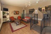 Located at 900 Country Club Dr SE in Rio Rancho, New Mexico, this single-floor community amenity space offers residents two distinct functional areas designed for relaxation and active living. The lounge area is bright and inviting, featuring warm hardwood-style flooring, large grid-pane windows that flood the room with natural light and provide views of the outdoor pool area. Comfortable wicker furnishings with rust-toned cushions, a glass-top coffee table, open shelving with decorative accents, and wall-mounted artwork create a casual yet welcoming atmosphere. The space also features a railing-lined overlook that connects to adjoining areas, adding an open, airy quality to the layout. The fitness center is a well-appointed workout space finished with durable carpet tile flooring and equipped with a variety of exercise machines, including a treadmill, elliptical, rowing machine, multi-station cable gym, and additional strength equipment. Floor-to-ceiling mirrors along one wall visually expand the room, while overhead fluorescent lighting ensures a well-lit environment. A secondary fitness area provides additional space for strength training with its own multi-function gym unit and natural light from a grid-pane window. Together, these amenities offer a functional and comfortable environment for the residents of this Rio Rancho community.