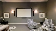 This sophisticated single-floor space is thoughtfully designed as a dedicated media and entertainment area, combining modern aesthetics with a functional layout. The centerpiece of the room is a large, wall-mounted projection screen flanked by sleek floor-standing speakers and framed by a pair of elegant wall sconces that cast a warm, ambient glow against the deep grey walls. A soundbar sits neatly beneath the screen, completing the high-quality audio-visual setup.

The interior design is cohesive and refined, with grey carpeting that complements the cool-toned wall color and contributes to the room's acoustic comfort. Stylish patterned accent chairs in grey and white provide comfortable seating while adding visual interest to the space. A glass-top side table and a black accent cube offer practical surfaces without disrupting the clean, open feel of the room.

Framed wall art featuring a serene seascape adds a touch of sophistication and rounds out the décor. The overall ambiance is intimate yet polished, striking a balance between a casual lounge and a purpose-built home theater. This space is ideal for immersive movie viewing, gaming, or simply relaxing in a well-appointed environment.