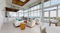 Located at 3680 46th Ave S in Saint Petersburg, Florida, this single-floor clubhouse and amenity space delivers an impressive coastal-modern experience defined by striking architectural design and abundant natural light. The centerpiece of the space is a grand lounge area with soaring double-height ceilings clad in warm wood paneling, anchored by expansive floor-to-ceiling glass curtain walls that frame sweeping panoramic views of the surrounding Saint Petersburg skyline and waterways. Large-format marble-look porcelain tile flooring runs throughout, reinforcing the clean, upscale aesthetic. The lounge is thoughtfully furnished with white upholstered sofas and accent chairs accented by teal throw pillows, natural wood side tables, and a statement chandelier, creating a sophisticated yet inviting atmosphere. A wraparound exterior terrace with glass railings extends the living space outdoors, offering unobstructed elevated views. The kitchen and service area features a sleek white bar counter, stainless steel appliances, globe pendant lighting, and a slatted wood accent wall. A separate high-top bar nook with a marble-top table, chrome stools, and a gold wall sconce provides an intimate setting. Restrooms and additional seating areas round out the floor plan, making this a fully equipped, visually stunning communal retreat.