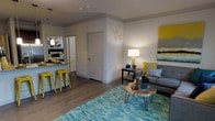 This well-appointed single-floor residence offers a thoughtfully designed open-concept layout that connects the living and kitchen areas in one cohesive, inviting space. The living area serves as the heart of the home, anchored by a plush gray sofa dressed with patterned and solid throw pillows in shades of yellow and blue. A striking teal area rug adds a vibrant focal point underfoot, while a natural wood slab coffee table on hairpin legs brings an organic, contemporary touch. A floor lamp and table lamp provide warm ambient lighting, complemented by colorful wall art that ties the color palette together. The kitchen features a breakfast bar with bright yellow metal stools, dark countertops with bold patterned accents, and a stainless steel refrigerator. Warm-toned hardwood-style flooring runs throughout the space, lending continuity and warmth. The walls are painted in a clean, neutral gray-white that amplifies the natural light and allows the décor accents to stand out. A paneled interior door leads to an adjacent room, suggesting a well-organized floor plan with defined private spaces. Overall, the residence balances modern design with vibrant, curated details for a lively yet functional living environment.