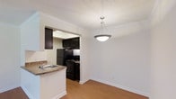 This single-floor, one-bedroom, one-bathroom unit offers a practical and well-organized layout designed for comfortable everyday living. The dining area is positioned adjacent to the kitchen and is defined by a contemporary pendant light fixture that hangs from a textured ceiling with crown molding detail, adding a refined touch to the space.

The kitchen is accessible via an open pass-through archway, creating a connected feel between the cooking and dining areas. It is outfitted with dark espresso cabinetry that provides ample storage, and features a full suite of black appliances, including a refrigerator, gas range, and additional built-in units. A countertop with an integrated sink sits at the pass-through, offering additional prep space and casual functionality.

Warm hardwood-style flooring runs throughout the main living areas, complementing the clean white walls and crisp white baseboards. The overall aesthetic is cohesive and neutral, creating a versatile backdrop. Crown molding accents the ceiling transition between rooms, lending a polished finish to the interior.