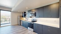 This thoughtfully designed one-bedroom, one-bathroom unit offers a modern living experience across a single well-appointed floor. The open-concept kitchen and living area serves as the heart of the home, featuring sleek dark cabinetry, light countertops, a full suite of stainless steel appliances including a refrigerator, electric range, microwave, and dishwasher, and light wood-style flooring that flows throughout the main living spaces. Large floor-to-ceiling sliding glass doors in the living room flood the space with natural light and provide direct access to a private balcony, where panoramic views of a city skyline and surrounding hills create a striking backdrop. A hallway with a stylish black-hardware barn door connects the living area to the bedroom and bathroom, and also houses a convenient stacked in-unit washer and dryer. The bedroom is a calm, carpeted retreat with a sliding closet and its own access to the balcony, offering continued city and mountain vistas. The bathroom features a clean, all-white tub-shower combination with matte black fixtures and a vanity with wood-toned cabinetry. Clean white walls and recessed lighting throughout contribute to the unit's bright, contemporary ambiance.