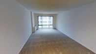 This single-floor space presents a clean, minimalist design centered around a spacious open living area. The room is finished with neutral wall-to-wall carpeting that adds warmth underfoot, while the crisp white walls and smooth ceiling create a bright, airy atmosphere throughout.

A prominent large sliding glass door with vertical blinds serves as the focal point of the main living area, allowing generous amounts of natural light to filter into the space. The door opens to an exterior view of a multi-story building facade, suggesting an urban setting with city surroundings visible beyond.

A compact radiator unit is positioned along one wall near the window, providing an integrated heating solution within the space. The overall layout is straightforward and open, offering a flexible arrangement with no obstructing columns or partitions. The simplicity of the design makes it adaptable for a variety of furniture configurations and personal uses.

The combination of soft carpeting, white walls, and ample natural light gives the space a neutral, versatile character that is well-suited for comfortable everyday living.