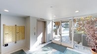 This building lobby offers a clean, well-organized entry experience designed for both practicality and ease of access. Upon entering through the glass-paneled double doors, natural light floods the space, illuminating the light-toned tile flooring and white ceiling fitted with recessed lighting. A wall-mounted bank of golden mailboxes lines the left side of the lobby, providing residents with convenient mail access directly at the entrance. Adjacent to the mailboxes, a smooth wood-finished elevator door offers vertical access within the building, accompanied by a nearby intercom or access control panel. The glass entry doors frame views of the outdoor surroundings, including a paved walkway, landscaping, and neighboring structures, giving the lobby an open, connected feel despite its compact footprint. A floor mat at the entrance adds a practical touch, helping maintain cleanliness within the space. The overall design is straightforward and modern, prioritizing function while maintaining a tidy, welcoming atmosphere for residents and visitors alike.