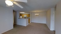 This single-floor space offers a functional and open-concept layout that seamlessly connects the kitchen, dining, and living areas into one cohesive living environment. The kitchen features warm honey-toned cabinetry, a dark countertop peninsula that doubles as a breakfast bar, and white appliances including a refrigerator and microwave. Under-cabinet fluorescent lighting provides practical task illumination in the kitchen. The peninsula creates a natural boundary between the kitchen and the main living area without closing off the space, maintaining an open and airy feel throughout. The dining area is accented by an elegant brass chandelier with candle-style lights, adding a classic decorative touch. The living area is finished with neutral wall-to-wall carpeting that lends warmth and comfort underfoot. A ceiling fan overhead in the living space ensures year-round climate comfort. The textured ceiling adds a subtle architectural detail consistent throughout the space. Neutral gray-white walls provide a clean, versatile backdrop suitable for a variety of interior styles. The layout is well-suited for comfortable everyday living, with each functional zone flowing naturally into the next.