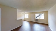 This single-floor space features a bright, open living area finished with rich, dark hardwood-style flooring that contrasts cleanly against the light beige walls. A textured popcorn ceiling adds a classic touch overhead, while a ceiling fan with an integrated light fixture provides both air circulation and ambient lighting. Two windows along the front wall allow ample natural light to fill the room, offering views of the exterior surroundings, including an outdoor staircase visible through one pane. A wall-mounted air conditioning unit sits beneath one of the windows, providing a dedicated climate control solution. A white entry door is centrally positioned between the windows, creating a straightforward and functional entryway. A partial wall with a pass-through counter separates the living area from an adjacent space, likely a kitchen, adding a sense of openness while maintaining a defined layout. White baseboards run along the perimeter, adding a clean, finished look. Overall, the space presents a functional and straightforward layout with practical features suited for everyday living.