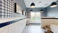 This thoughtfully designed pet grooming space makes a strong visual impression with its signature navy and white houndstooth-patterned upper walls, which are complemented by crisp white subway tile wainscoting trimmed with bold navy blue accent borders. The light-toned large-format floor tiles keep the space feeling clean and bright, while a floor drain ensures practical functionality. A built-in deep wash basin with a faucet is elevated on a stepped platform, making it easily accessible for bathing pets of various sizes. The steps leading up to the basin are finished in a neutral tone, blending seamlessly with the overall design. A row of white open-cube shelving units provides organized storage for grooming supplies and accessories. Multiple black pendant lights hang from the ceiling, delivering focused illumination throughout the room. A large window along one wall floods the space with natural light and offers a pleasant view of the surrounding greenery. Framed pet-themed artwork adorns the walls, adding a playful and welcoming character to the space. A glass-paneled door provides access to an adjacent corridor, contributing to an open, connected feel. Overall, the space combines stylish aesthetics with highly practical, purpose-built features for a premium pet grooming experience.