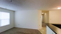 This single-floor residence presents a straightforward and functional layout. The main living area is finished with wall-to-wall carpeting in a neutral tan tone, providing a warm underfoot feel. Smooth white walls and a textured popcorn ceiling define the space, contributing to a clean, understated aesthetic throughout.

A partial wall divides the main living area from an adjoining transition zone, where the flooring shifts to light-colored tile, suggesting proximity to a kitchen or entryway. A countertop edge is partially visible in the foreground, indicating a kitchen or bar area adjacent to the main living space.

A hallway extends toward the back of the unit, illuminated by a flush-mount ceiling light, and leads to what appear to be additional rooms, likely bedrooms and a bathroom. Doors along the hallway suggest multiple private spaces branching off this central corridor.

The overall color palette is neutral and consistent, with beige tones dominating the walls, flooring, and ceiling. The unit appears well-maintained and move-in ready, offering a blank canvas suited for a variety of furnishing styles.