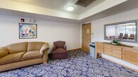 Located at 770 Savanna Ave in Saint Cloud, Minnesota, this single-floor community room is a well-appointed, multipurpose space designed to accommodate a variety of gatherings and activities. The room is thoughtfully divided into distinct functional zones, offering both a dining and meeting area as well as a comfortable lounge section.

The dining area is anchored by a large rectangular wooden table surrounded by matching wood-slat chairs, providing ample seating for group meals or organized meetings. Adjacent to this area, a lounge zone features upholstered armchairs and a sofa, creating a relaxed setting for casual conversation or smaller gatherings. Additional stacked banquet chairs stored near the window offer flexibility for expanding seating capacity when needed.

A large window along one wall allows natural light to fill the space while offering views of the surrounding outdoor area. The textured ceiling and flush-mount lighting fixtures provide consistent, even illumination throughout the room. The distinctive blue patterned carpet adds a warm, welcoming character to the interior, complementing the neutral-toned walls.

A wooden credenza with a small refrigerator and a decorative mirror are situated near the entry door, adding a practical and finished touch. A mechanical room door is also accessible from within the space. Overall, this community room offers a functional and inviting environment well-suited for social events, meetings, and shared activities.