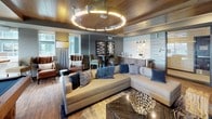 This contemporary entertainment lounge is thoughtfully designed to deliver a premium social experience across a single open floor. The space is anchored by a full-size billiards table set atop warm hardwood flooring, complemented by a generous seating area featuring a large sectional sofa, leather armchairs, and a bold coffee table with an organic sculptural centerpiece. A dramatic circular chandelier with warm LED lighting serves as a striking focal point overhead, while a rich wood-paneled ceiling adds depth and a refined, lodge-inspired warmth throughout.

One wall showcases a textured, wave-patterned accent panel in a cool metallic tone, providing a sleek backdrop for a large wall-mounted flat-screen television. A set of velvet blue cube ottomans completes this media viewing zone with casual, stylish seating. On the opposite end, a wet bar with white subway tile, modern pendant lighting, and wood cabinetry offers a polished serving station ideal for gatherings.

Large sliding glass doors flood the billiards area with natural light and provide direct access to an outdoor terrace, seamlessly connecting the interior to the exterior. An entry vestibule with glass-paneled doors and privacy film adds a refined transition point into the lounge. The overall design balances modern sophistication with inviting comfort, making it a well-appointed space for recreation and social events.