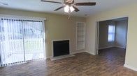 This single-floor space offers a well-appointed layout designed for comfortable everyday living. The main living area serves as the heart of the home, showcasing rich, dark hardwood-style flooring that runs throughout and adds warmth to the space. A classic fireplace anchors the room, providing a natural focal point and a cozy atmosphere. Adjacent to the fireplace, a set of built-in shelves offers convenient storage and display space, adding both function and architectural character to the room. A ceiling fan with an integrated light fixture is centrally mounted overhead, ensuring year-round comfort and efficient air circulation. Natural light fills the room through a large sliding glass door dressed with vertical blinds, which opens to an outdoor area and creates a seamless connection between interior and exterior living. The neutral, warm-toned walls complement the flooring and contribute to a bright, inviting ambiance throughout. An additional doorway leads to further rooms within the space, hinting at a thoughtful and practical floor plan. The textured ceiling adds a subtle design detail that completes the overall character of this well-maintained, move-in-ready space.