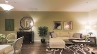 This single-floor space presents a thoughtfully designed living area that balances comfort and style. The room is defined by warm sage green walls that lend a calm, inviting atmosphere, complemented by rich dark hardwood flooring that anchors the space with a sense of warmth and depth.

The living area is furnished with a generous cream-toned sectional sofa accented with patterned throw pillows in navy and white, positioned atop a bold, dark-toned area rug featuring an intricate scrollwork design. A low-profile dark coffee table completes the seating arrangement, adding contrast to the lighter furnishings.

Along one wall, a dark wood console or bar cabinet provides functional storage and display space, topped with a decorative table lamp. Adjacent to it, a striking sunburst-style round mirror with a metallic finish serves as an eye-catching focal point. Framed botanical and still-life artwork arranged in a pair adds visual interest to another wall section.

A lush indoor potted tree introduces a natural, organic element to the room, softening the overall aesthetic. Crown molding along the ceiling adds a refined architectural detail, contributing to the polished, well-appointed feel of the space.