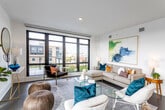 This contemporary single-floor residence is defined by its striking floor-to-ceiling black-framed windows, which flood the open living area with natural light and frame sweeping urban views of the surrounding cityscape. The spacious living room offers a versatile layout, accommodating multiple seating arrangements around a clear acrylic coffee table, with crisp white walls providing an ideal backdrop for bold artwork and eclectic décor accents.

The kitchen is thoughtfully designed with high-gloss white upper cabinetry, warm wood-toned lower cabinets, and a generous island with a clean white countertop and bar seating. Stainless steel double wall ovens and a gas cooktop underscore the space's culinary functionality, while wide-plank hardwood flooring adds warmth throughout.

The primary bedroom is a serene retreat featuring dark hardwood floors, a large window with urban views, and refined furnishings complemented by curated artwork. A second bedroom offers built-in closets, recessed lighting, and a calm, neutral palette. The primary bathroom is spa-inspired, with large-format tiles, a glass-enclosed walk-in shower, dual vanities with light wood cabinetry, and white quartz countertops. A separate powder room provides convenience for guests, finished with a floating vanity and soft, muted tones throughout.