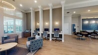 This single-floor community clubhouse is thoughtfully designed to serve multiple functions within a cohesive, elegantly finished space. The main lounge area features light hardwood flooring, soaring ceilings with crown molding, and large windows that flood the room with natural light and offer outdoor views. A modern circular chandelier anchors the seating area, which is furnished with patterned accent chairs and a central coffee table atop a geometric area rug.

Adjacent to the lounge, the business center offers a row of individual computer workstations built into a continuous desk, separated by white decorative columns with wall sconces providing focused task lighting. Ergonomic task chairs and desktop computers make this a fully functional workspace.

A private conference room features steel-blue walls adorned with framed artwork, carpeted flooring, a round wooden table surrounded by contemporary upholstered chairs, and a wall-mounted television, making it well-suited for meetings or presentations.

The dining area provides multiple seating configurations, including round tables with curved chairs and counter-height bar seating, illuminated by globe pendant lights. Natural light pours in through large windows dressed with blinds and floor-length drapes, completing the refined, welcoming ambiance throughout.