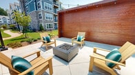 Located at 640 Lakeside Dr in Canton, GA, this outdoor lounge area serves as a welcoming community gathering space designed for relaxation and social connection. The patio is anchored by a sleek, square fire pit finished in a dark charcoal tone, surrounded by a generous arrangement of natural wood Adirondack-style chairs accented with teal cushions, creating a comfortable and inviting atmosphere. The space is defined by striking warm-toned horizontal wood plank accent walls that lend a sense of enclosure and privacy while adding rich, natural texture to the design. Overhead, strands of globe string lights are suspended between black poles, providing ambient evening illumination and a festive character. The concrete patio surface is laid in large-format tiles, offering a clean and polished foundation throughout the seating area. Lush landscaping with manicured greenery, young trees, and mulched beds borders the space, softening the hardscape and integrating the lounge into the broader community grounds. Views from the patio extend to well-maintained lawns, walking paths, and the multi-story residential buildings of the surrounding community, reinforcing its role as a central outdoor amenity. The open-sky setting ensures ample natural light, making this a versatile space for daytime lounging and evening gatherings alike.