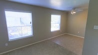 This single-story space features a straightforward, functional layout highlighted by neutral design choices throughout. The main living area is finished with wall-to-wall carpeting in a beige tone, complementing the taupe-painted walls and white baseboards that frame the room cleanly. Two windows fitted with horizontal blinds allow natural light to filter into the space, offering views of the surrounding exterior. A ceiling fan with a light fixture is mounted overhead, providing both ventilation and ambient lighting. The textured ceiling adds subtle visual depth to the room. The overall ambiance is calm and understated, presenting a blank canvas well-suited for a variety of furniture arrangements and interior styles.
