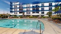 Located at 13900 Tahiti Way in Marina del Rey, California, this outdoor amenity space delivers a resort-style experience defined by thoughtful design and a prime coastal setting. The centerpiece is a generously sized swimming pool with sparkling blue water, framed by wide concrete pool decking that accommodates rows of wooden-framed chaise lounges paired with large navy umbrellas — ideal for lounging under the Southern California sun. A separate spa or wading area is positioned adjacent to the main pool, adding to the relaxation offerings. The surrounding landscape is carefully curated, featuring lush shrubs, flowering plants, ornamental grasses, and mature trees that provide both privacy and natural beauty. A shade structure with a distinctive latticed canopy offers a sheltered retreat within the outdoor space. The backdrop is dominated by the building's bold, undulating architectural facade — white curved forms accented with vivid blue wave-shaped glass balcony panels that evoke the nearby Pacific Ocean. Views beyond the perimeter fence reveal the masts of sailboats docked at the marina, reinforcing the waterfront character of this location. The overall ambiance is that of a well-appointed, design-forward outdoor retreat in one of Los Angeles County's most sought-after coastal communities.