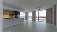This contemporary single-floor space showcases a thoughtfully designed open-concept layout that seamlessly integrates the living area and kitchen. Expansive floor-to-ceiling windows wrap the living space, flooding the interior with natural light while offering sweeping views of the surrounding cityscape and treetops beyond. The light-toned hardwood flooring extends throughout, enhancing the sense of openness and continuity across the space.

The kitchen is positioned along one side of the open floor plan and features dark cabinetry that provides a striking contrast to the lighter tones of the living area. Stainless steel appliances, including a refrigerator and microwave, contribute to the modern aesthetic, while under-cabinet lighting adds warmth to the workspace. A kitchen counter peninsula serves as a natural divider between the culinary and living zones.

The living area is generously proportioned, offering ample room for a variety of furnishing arrangements. A ceiling fan is centrally mounted, providing both functional air circulation and a subtle design element. Horizontal window blinds offer privacy and light control without compromising the connection to the outdoors. Overall, the space exudes a clean, contemporary ambiance defined by natural light, modern finishes, and an airy, connected layout.