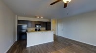 This single-floor residence showcases an open-concept layout that integrates the kitchen and living area into one cohesive, functional space. The kitchen is outfitted with rich, dark wood cabinetry that provides ample storage, complemented by granite countertops featuring warm tones. A peninsula with bar-height seating extends from the kitchen, creating a natural division between the cooking and living spaces while maintaining an open feel. Black appliances, including a refrigerator, microwave, and range, are built seamlessly into the cabinetry. Track lighting above the kitchen ensures well-lit food preparation areas.

The living area extends generously from the kitchen, finished with durable hardwood-style flooring that runs throughout the open space, adding warmth and continuity. A ceiling fan with an integrated light fixture is centrally positioned overhead, promoting air circulation and supplemental lighting. Neutral-toned walls throughout keep the space feeling bright and versatile. A white paneled door near the back of the living area leads to additional rooms, suggesting a well-organized, practical floor plan designed for comfortable everyday living.