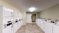 This shared laundry facility is a practical, single-floor space designed to efficiently serve multiple residents. The room is outfitted with a row of top-loading, coin-operated washing machines aligned along one wall, providing ample washing capacity. Opposite the washers, a bank of stacked coin-operated dryers maximizes the use of vertical space, offering an equal number of drying units to match laundry demand. A utility sink with cabinetry is conveniently located at one end of the washer row, ideal for pre-treating garments or hand-washing delicate items. The space also includes a small table and seating area, allowing residents to wait comfortably while their laundry cycles complete. A bulletin board on the wall provides a place for community notices and laundry instructions. The room is finished with neutral-toned walls and large-format ceramic tile flooring that is easy to maintain and clean. Overhead flush-mount lighting ensures the space is well-lit throughout. Safety is addressed with a clearly mounted fire extinguisher. Access to the room is provided through a standard door, with additional corridor access visible, connecting the laundry room to the broader building.