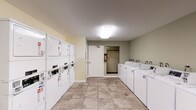 This shared laundry facility is a practical, single-floor space designed to efficiently serve multiple residents. The room is outfitted with a row of top-loading, coin-operated washing machines aligned along one wall, providing ample washing capacity. Opposite the washers, a bank of stacked coin-operated dryers maximizes the use of vertical space, offering an equal number of drying units to match laundry demand. A utility sink with cabinetry is conveniently located at one end of the washer row, ideal for pre-treating garments or hand-washing delicate items. The space also includes a small table and seating area, allowing residents to wait comfortably while their laundry cycles complete. A bulletin board on the wall provides a place for community notices and laundry instructions. The room is finished with neutral-toned walls and large-format ceramic tile flooring that is easy to maintain and clean. Overhead flush-mount lighting ensures the space is well-lit throughout. Safety is addressed with a clearly mounted fire extinguisher. Access to the room is provided through a standard door, with additional corridor access visible, connecting the laundry room to the broader building.