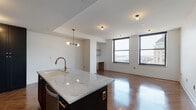 This thoughtfully designed high-rise residence combines contemporary finishes with an open, flowing layout spread across a single level. The kitchen serves as the centerpiece of the living area, showcasing a large island with a striking marble countertop, a brass gooseneck faucet, and bold dark cabinetry that reaches the ceiling. Stainless steel appliances, a sleek range hood, and a marble tile backsplash complete the chef-quality space. A modern globe chandelier adds an elegant touch above the island. Rich herringbone-patterned hardwood floors run throughout the entire residence, lending warmth and a refined aesthetic to every room. The open living area flows naturally from the kitchen and is framed by large, black-trimmed windows that frame sweeping city skyline and water views, flooding the space with natural light. The bedrooms continue the high-end feel, with corner window configurations that maximize the panoramic urban vistas. The bathrooms are finished with crisp white subway tile, natural wood vanities with marble countertops, matte black fixtures, and warm brass accent lighting. Built-in shelving and thoughtful nooks add functional detail throughout. The overall ambiance is clean, modern, and sophisticated, with a consistent palette of warm wood, brass, and neutral tones.