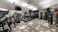 This fitness center is thoughtfully designed to accommodate a wide range of workout styles within a single, open floor plan. The space is finished with a distinctive patterned carpet in neutral grey and white tones, lending a modern aesthetic throughout. Overhead, recessed and linear fluorescent lighting ensures the entire area is well-illuminated, complemented by ceiling-mounted speakers and security equipment.

The cardio zone features a row of Matrix treadmills and elliptical machines, each equipped with individual digital displays. Stair climbers and stationary bikes round out the cardio offerings. Multiple wall-mounted flat-screen televisions are positioned throughout for entertainment during workouts.

A dedicated strength training area includes a tiered dumbbell rack with a full range of free weights, paired with Matrix adjustable benches. A cable functional trainer provides versatility for resistance-based exercises. The functional training corner is equipped with a TRX suspension system, a punching bag, medicine balls, kettlebells, and a selection of stability and exercise balls stored on dedicated racks.

Full-length mirrors line one wall, enhancing the sense of space. A frosted glass partition defines the entry zone, while a water cooler provides convenient hydration. The overall ambiance is clean, professional, and well-organized.