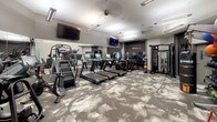 This fitness center is thoughtfully designed to accommodate a wide range of workout styles within a single, open floor plan. The space is finished with a distinctive patterned carpet in neutral grey and white tones, lending a modern aesthetic throughout. Overhead, recessed and linear fluorescent lighting ensures the entire area is well-illuminated, complemented by ceiling-mounted speakers and security equipment.

The cardio zone features a row of Matrix treadmills and elliptical machines, each equipped with individual digital displays. Stair climbers and stationary bikes round out the cardio offerings. Multiple wall-mounted flat-screen televisions are positioned throughout for entertainment during workouts.

A dedicated strength training area includes a tiered dumbbell rack with a full range of free weights, paired with Matrix adjustable benches. A cable functional trainer provides versatility for resistance-based exercises. The functional training corner is equipped with a TRX suspension system, a punching bag, medicine balls, kettlebells, and a selection of stability and exercise balls stored on dedicated racks.

Full-length mirrors line one wall, enhancing the sense of space. A frosted glass partition defines the entry zone, while a water cooler provides convenient hydration. The overall ambiance is clean, professional, and well-organized.