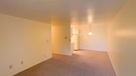 This single-floor residential space offers a straightforward and functional layout designed for comfortable everyday living. The open living area is finished with neutral-toned wall-to-wall carpeting that runs throughout, creating a cohesive and warm atmosphere across the main living space. Smooth, light-colored walls and a standard ceiling height contribute to a clean, understated aesthetic throughout.

A partial dividing wall separates the living area from the kitchen zone, providing a sense of defined space while maintaining an open feel between the two areas. The kitchen features white upper cabinetry and a white refrigerator, lending a bright and tidy appearance to the cooking space. A pendant light fixture hangs above, providing focused illumination and adding a modest decorative touch. The kitchen area transitions naturally into the main living space, making the overall floor plan feel connected and easy to navigate.

Recessed lighting in the living area provides additional overhead illumination, complementing the ambient warmth of the space. Multiple electrical outlets are positioned along the walls, offering practical convenience throughout. The overall ambiance is calm and neutral, making it a versatile space well-suited for a variety of furnishing and decorating styles.
