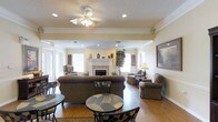 This single-story space combines comfortable interior living areas with well-appointed outdoor amenities. The interior lounge area is anchored by a classic white fireplace mantel, which serves as a focal point beneath a textured ceiling adorned with recessed lighting and a ceiling fan. Crown molding and chair rail detailing along the walls add a traditional architectural touch, while warm hardwood flooring runs throughout. The room is furnished with plush seating arrangements, including a large sofa, wingback chairs, and upholstered accent chairs, creating an inviting gathering environment. Decorative elements such as framed artwork, a tall potted plant, and mantel accessories contribute to the refined yet approachable ambiance. Natural light filters in through multiple windows fitted with blinds, brightening the neutral, cream-toned walls. Bistro-style tables with mosaic tops provide casual seating near the entrance. Outside, a generously sized swimming pool is enclosed by a wrought iron fence and flanked by white lounge chairs on a concrete pool deck. The brick exterior building and surrounding mature trees create a serene, park-like setting ideal for outdoor relaxation.