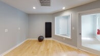 This single-level fitness studio is thoughtfully designed to support a variety of workout and wellness activities. The space is anchored by smooth hardwood flooring in a warm natural finish, providing an ideal surface for yoga, stretching, and bodyweight training. Multiple dark exercise mats are arranged across the floor, accommodating several participants simultaneously.

A large floor-to-ceiling mirror spans one full wall, visually expanding the room and allowing practitioners to monitor their form during exercise. Two wall-mounted flat-screen televisions are positioned for clear viewing from anywhere in the studio, complemented by a smaller interactive display panel—ideal for streaming guided workouts or fitness programs. Recessed ceiling lights provide bright, even illumination throughout the space.

The walls are finished in a clean, neutral gray tone, contributing to a calm and focused atmosphere. Additional fitness equipment, including a medicine ball and a rolled exercise mat, is stored neatly along the perimeter. An exterior door provides direct access, adding convenience for instructors and participants. The overall design is clean, functional, and purposefully minimalist, creating an environment well-suited for both individual training and small group fitness sessions.