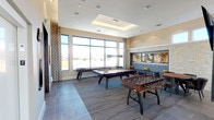 This contemporary amenity space is thoughtfully designed to offer a full range of social and recreational experiences within a single, well-appointed floor. The open layout is unified by large-format tile flooring and warm neutral tones, creating a cohesive and inviting atmosphere throughout.

The game room area is a standout feature, equipped with a billiards table and foosball table, set atop a textured area rug beneath a coffered ceiling with recessed lighting. Oversized windows flood the space with natural light and frame open outdoor views, complemented by elegant geometric-patterned drapes.

A sophisticated lounge nook offers a built-in banquette upholstered in sleek black leather, accented with bold diamond-pattern throw pillows. Above it, a natural stone accent wall frames a triptych of abstract artwork, adding a gallery-quality focal point to the seating area.

The wet bar and entertainment zone is anchored by a dramatic stone-veined marble island beneath a row of sculptural pendant lights. Dark cabinetry, a mosaic tile backsplash, and dual wall-mounted flat-screen televisions lend the bar a modern, high-end club aesthetic. A deep undermount sink and integrated appliances complete the functional setup.

Corner windows bring in abundant sunlight and sweeping exterior views, enhancing the open, airy character of the entire space.